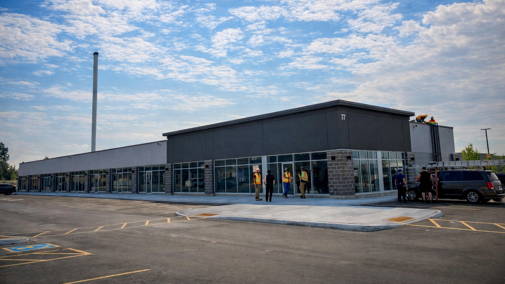 A single‑storey commercial building with a dark gray facade and large glass storefronts lines a wide parking area. Several people stand near the entrance under a partly cloudy blue sky.